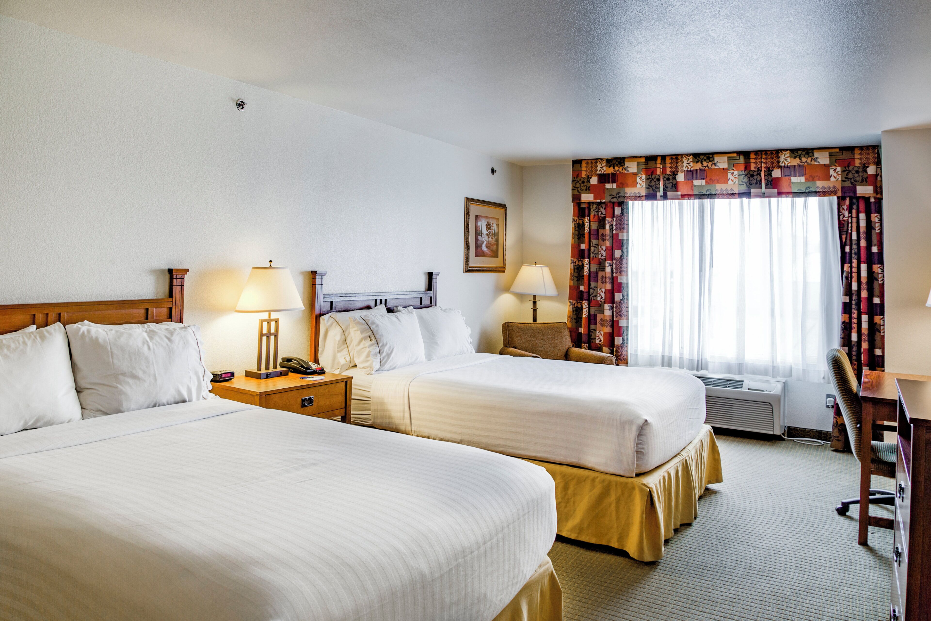 Habitación de hotel con dos camas, lámparas de noche, mesitas de noche, un escritorio, una silla, un ventanal con cortinas y un cuadro en la pared.