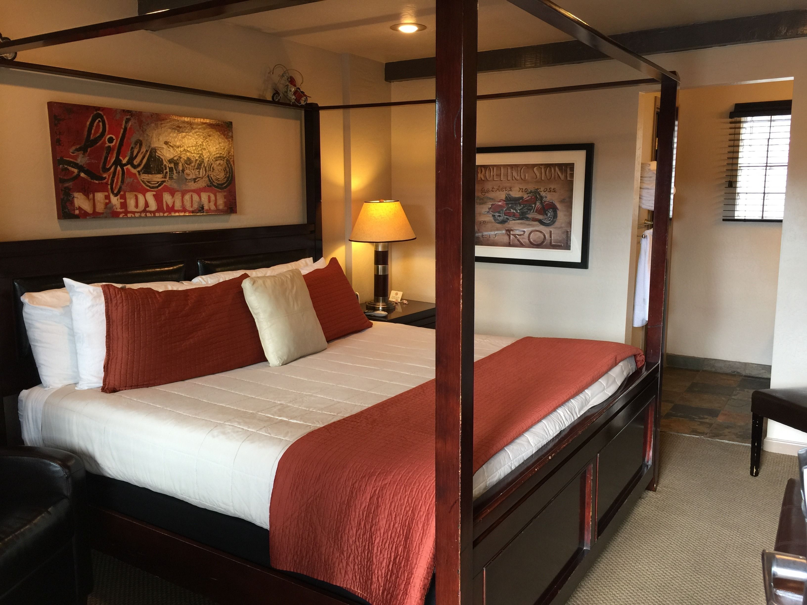 A four-poster bed with a red and white bedspread, a headboard with a framed picture, a bedside table with a lamp, and a framed picture on the wall.