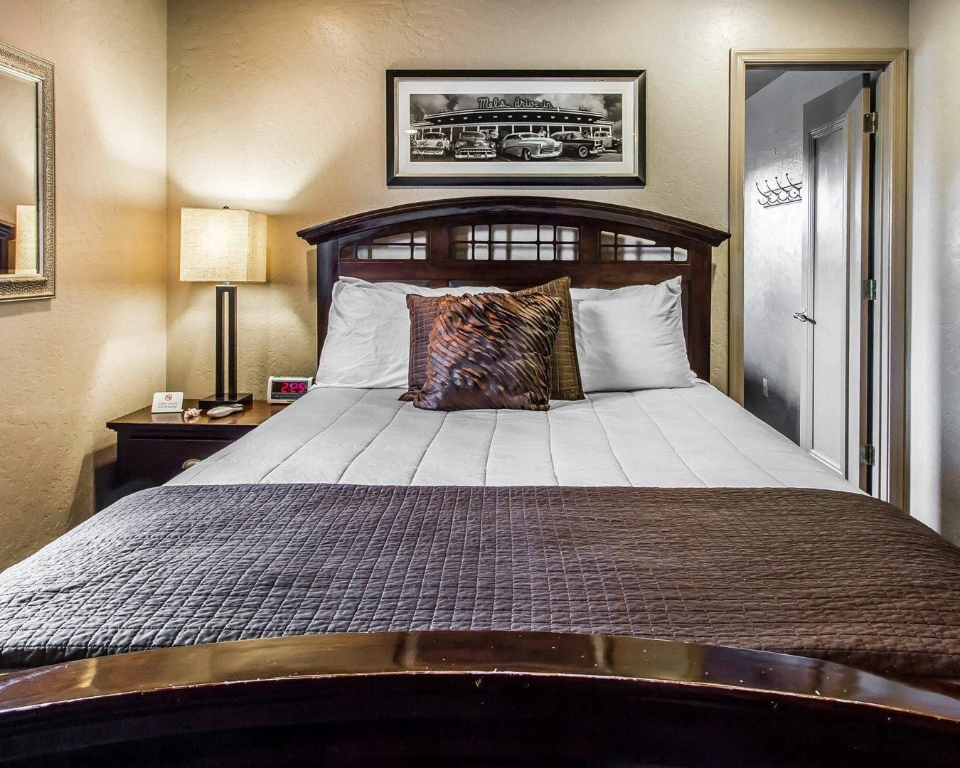 Et hotelværelse med en seng, et natbord med en lampe, et billede i ramme på væggen og en dør med et skilt.