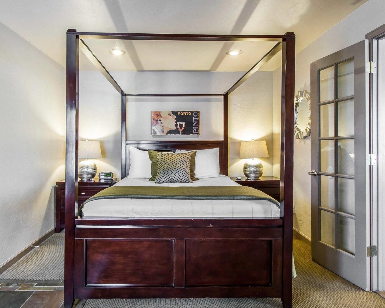 A four-poster bed with a headboard, two bedside tables with lamps, a framed artwork above the bed, and a door with glass panels to the right.