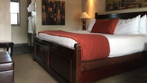 A hotel room with a large bed, a nightstand, a lamp, a painting on the wall, and a window with blinds. - Rodeway Inn & Suites Williams Downtowner - Rte 66 (Williams)