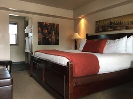Habitación de hotel con una cama grande, una mesita de noche, una lámpara, un cuadro en la pared y una ventana con persianas.