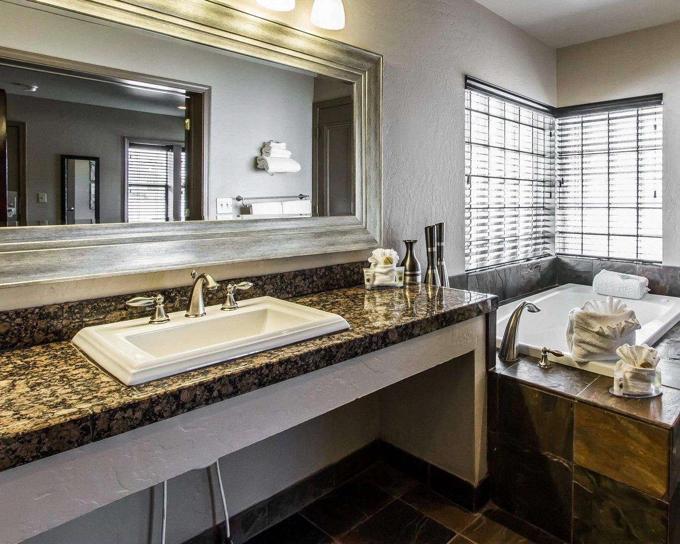 A modern bathroom with a granite countertop, a large mirror, a sink, a bathtub, and a window with blinds.