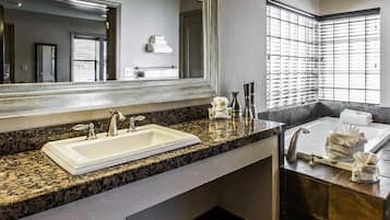 A modern bathroom with a granite countertop, a large mirror, a sink, a bathtub, and a window with blinds.