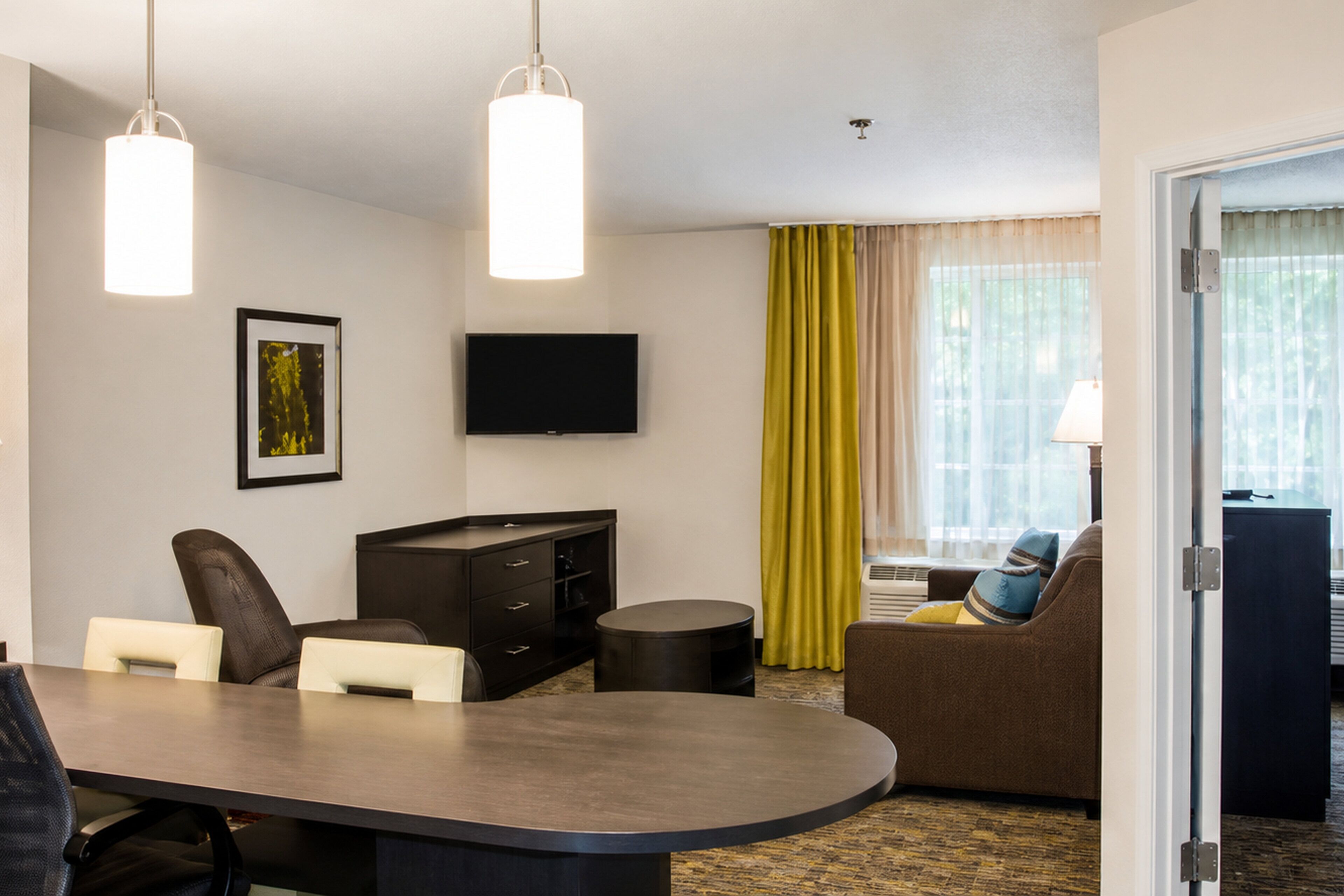 A hotel room with a dining table, chairs, a sofa, a TV, and a window with curtains.