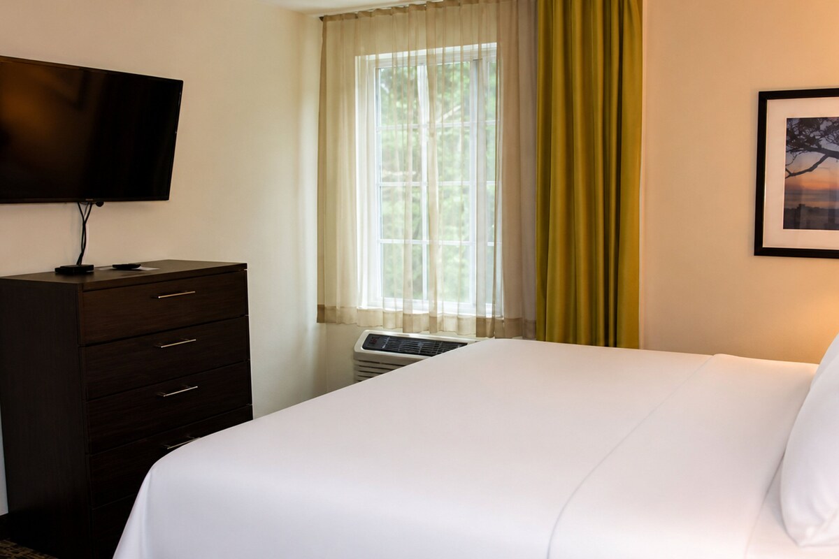 Een hotelkamer met een bed, een televisie op een dressoir, een raam met gordijnen en een lijst met een afbeelding aan de muur.