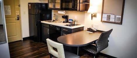 A compact kitchen with a refrigerator, microwave, and coffee maker. There is a dining table with two chairs and a lamp.