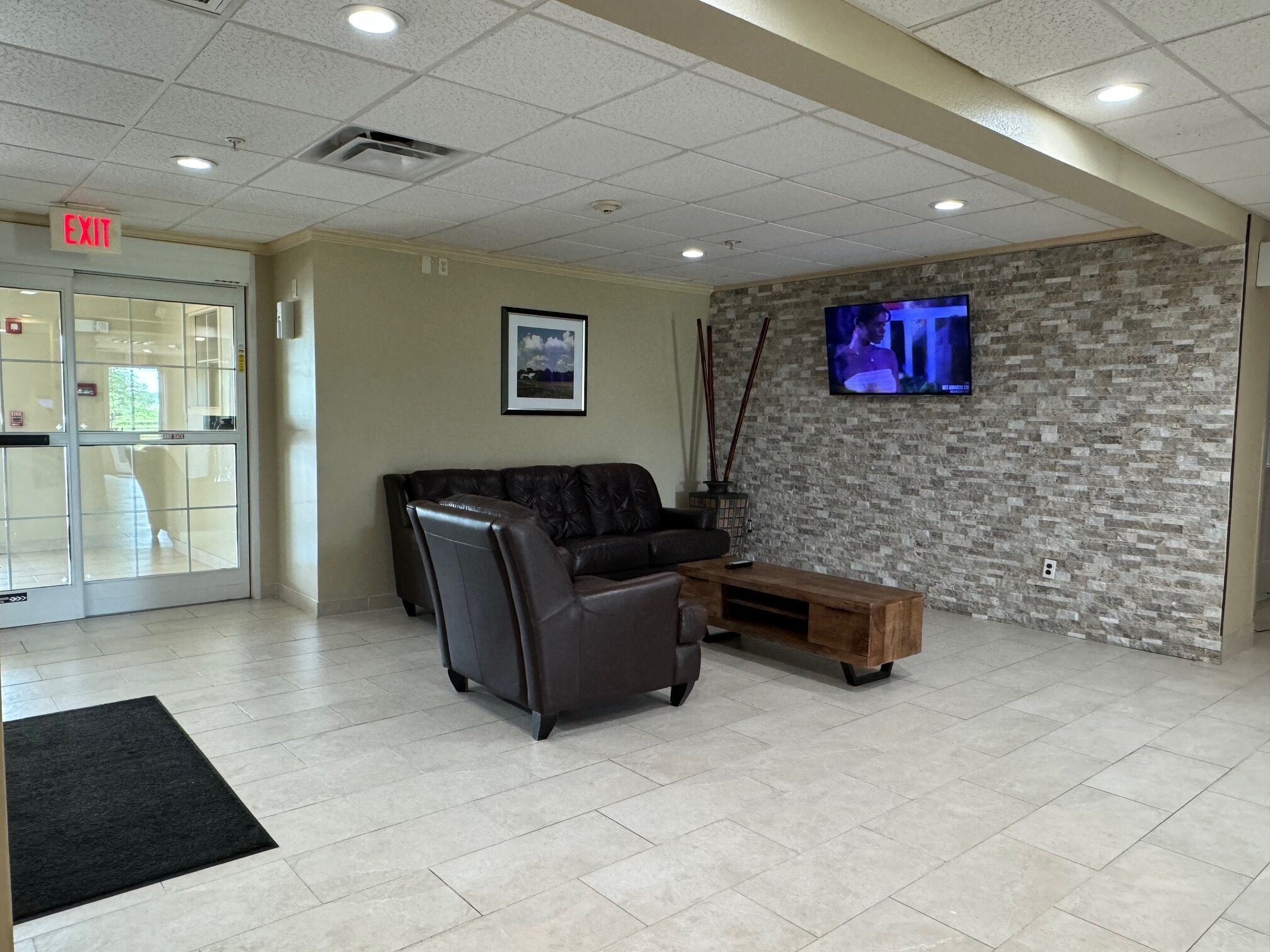 A lobby with a leather sofa, a wooden coffee table, a stone wall, a television, and an exit sign.