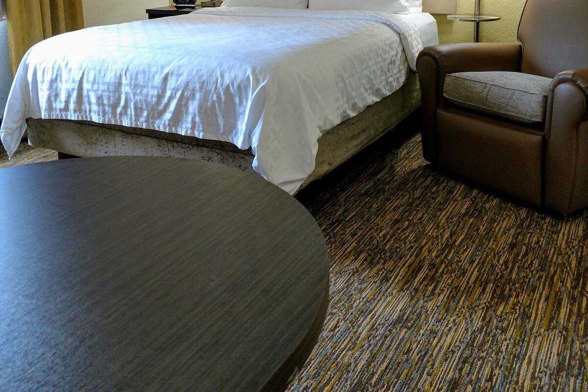 A hotel room with a bed, a chair, a table, and a lamp.
