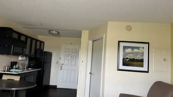 Una cocina compacta con armarios oscuros, un refrigerador y una cafetera. Hay una zona de comedor con una mesa y una silla. Una puerta conduce a otra habitación, y hay un cuadro en la pared.