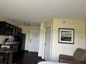 A compact kitchen with dark cabinets, a refrigerator, and a coffee maker. There is a dining area with a table and a chair. A door leads to another room, and a framed picture is on the wall. - Candlewood Suites Olive Branch by IHG (Olive Branch)