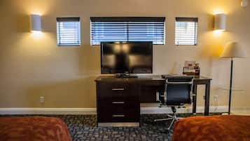 A hotel room with a desk, two chairs, a TV, and two windows with blinds.