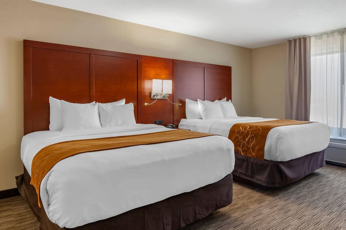 A hotel room with two beds, a wooden headboard, a wall-mounted light, and a window with curtains.