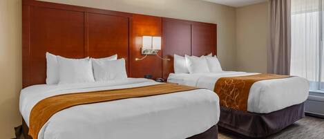 A hotel room with two beds, a wooden headboard, a wall-mounted light, and a window with curtains.