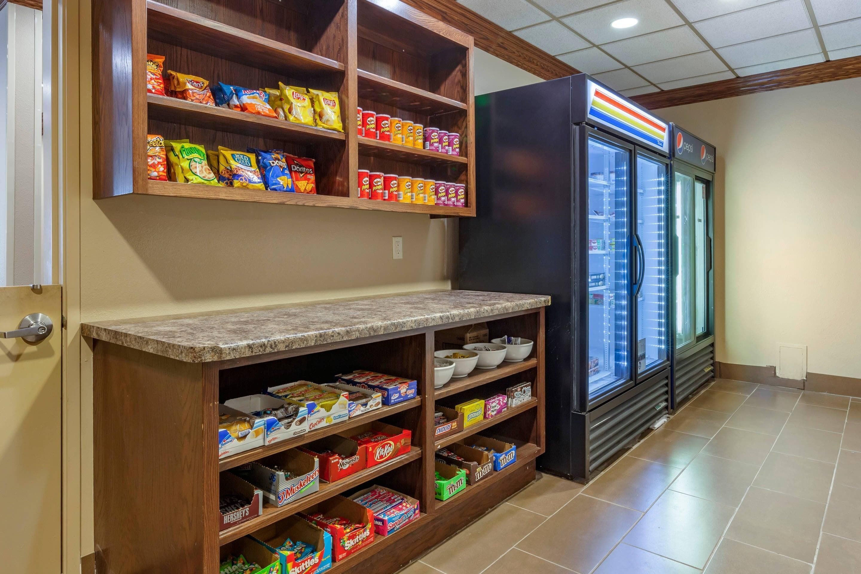A kitchen area with a refrigerator, shelves stocked with snacks, and a tiled floor.