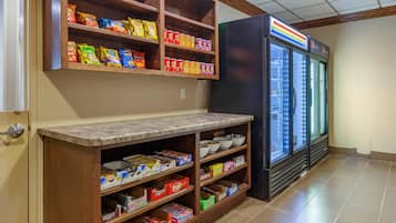A kitchen area with a refrigerator, shelves stocked with snacks, and a tiled floor.