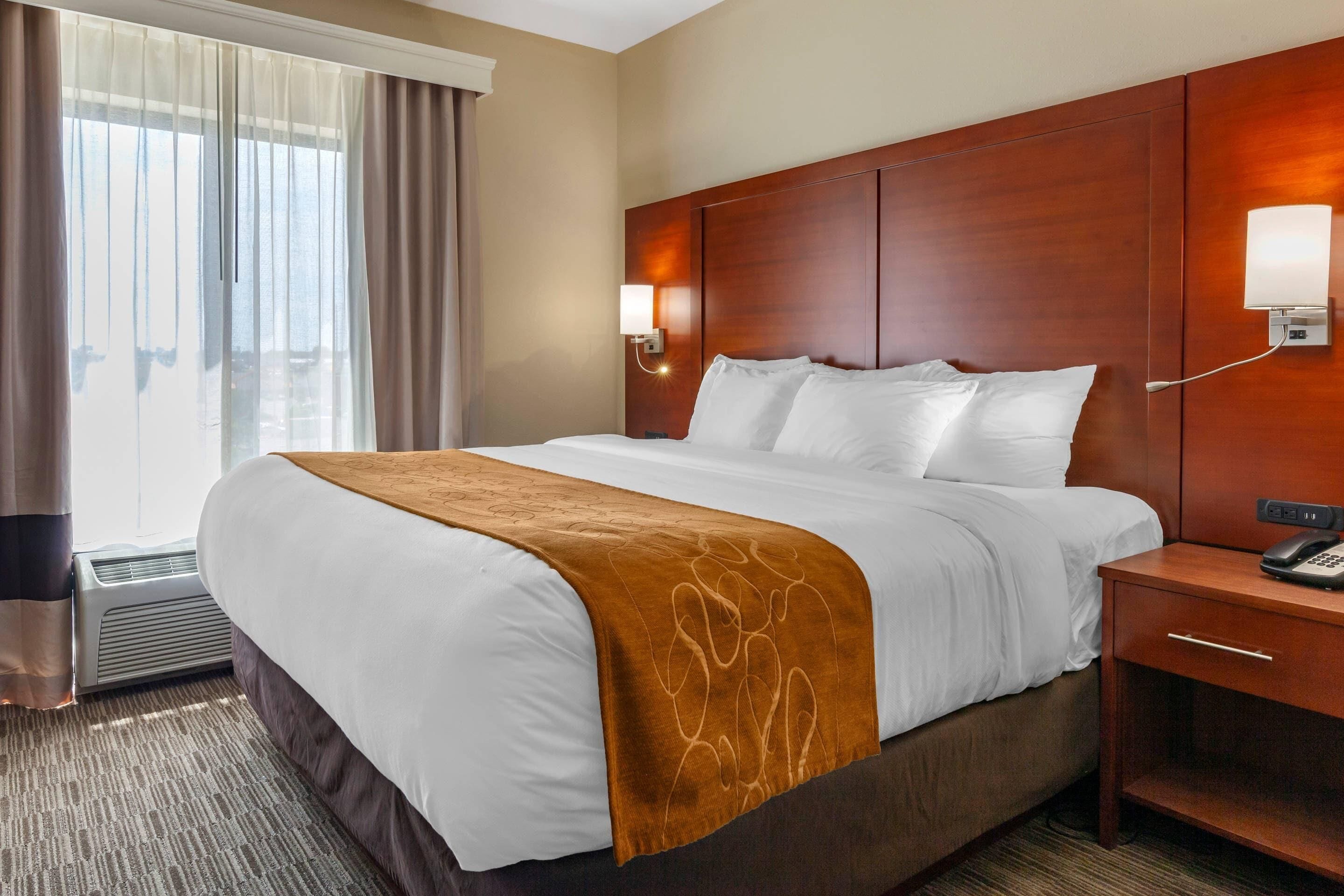 A hotel room with a large bed, a wooden headboard, a bedside table with a phone, and a window with curtains.