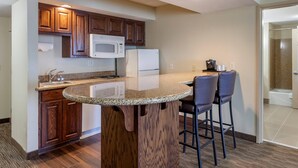 A kitchen with wooden cabinets, a microwave, a refrigerator, and a bar area with two stools. - Comfort Suites Oshkosh West (Oshkosh)