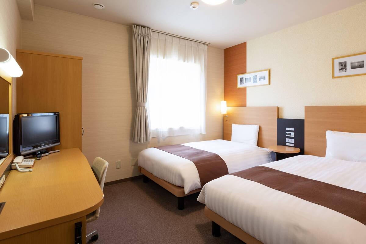 Een hotelkamer met twee bedden, een bureau met een computer, een stoel en een raam met gordijnen.