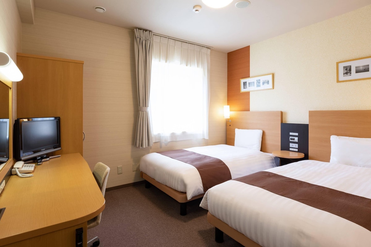 A hotel room with two beds, a desk with a computer, a chair, and a window with curtains.