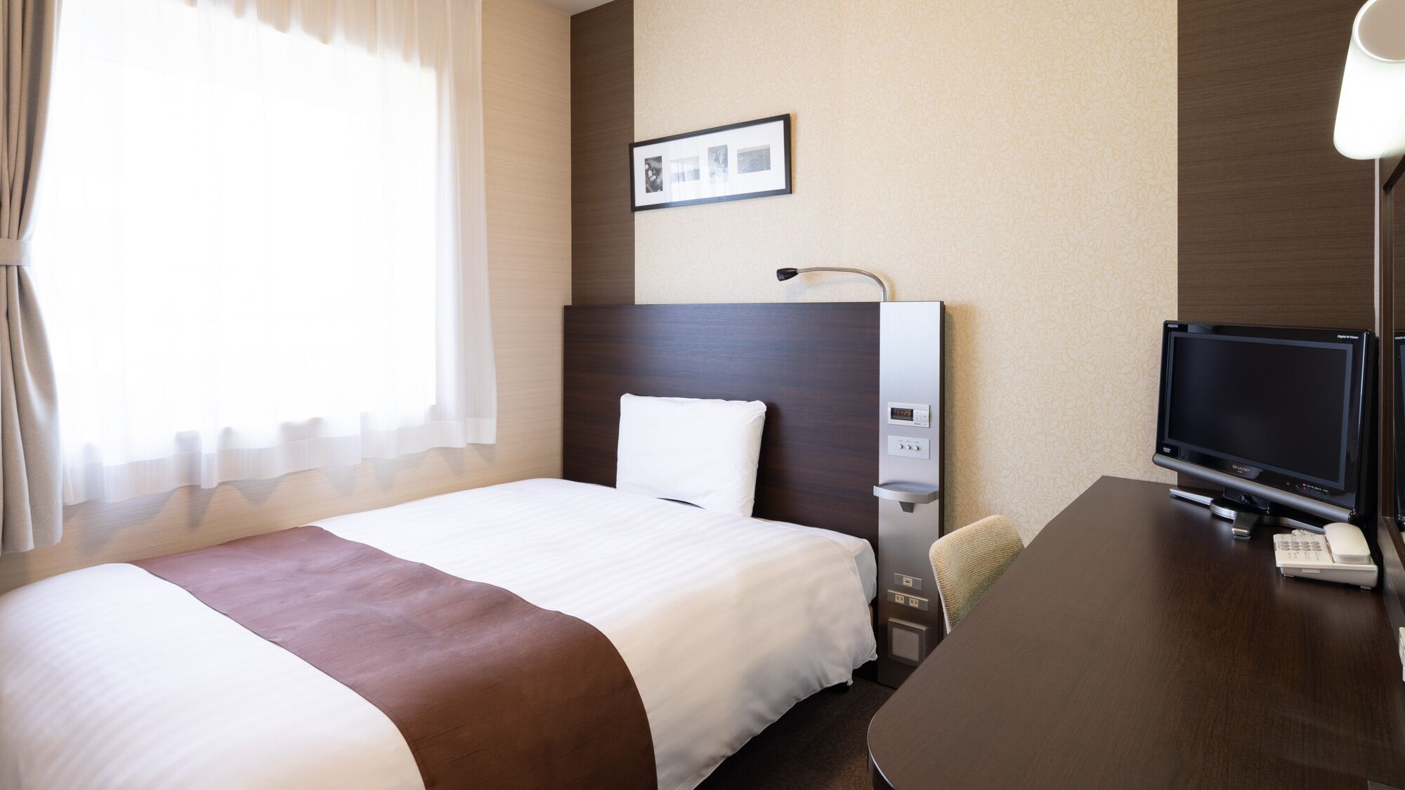 A hotel room with a bed, a desk with a TV, a desk lamp, a wall-mounted picture, and a window with curtains.