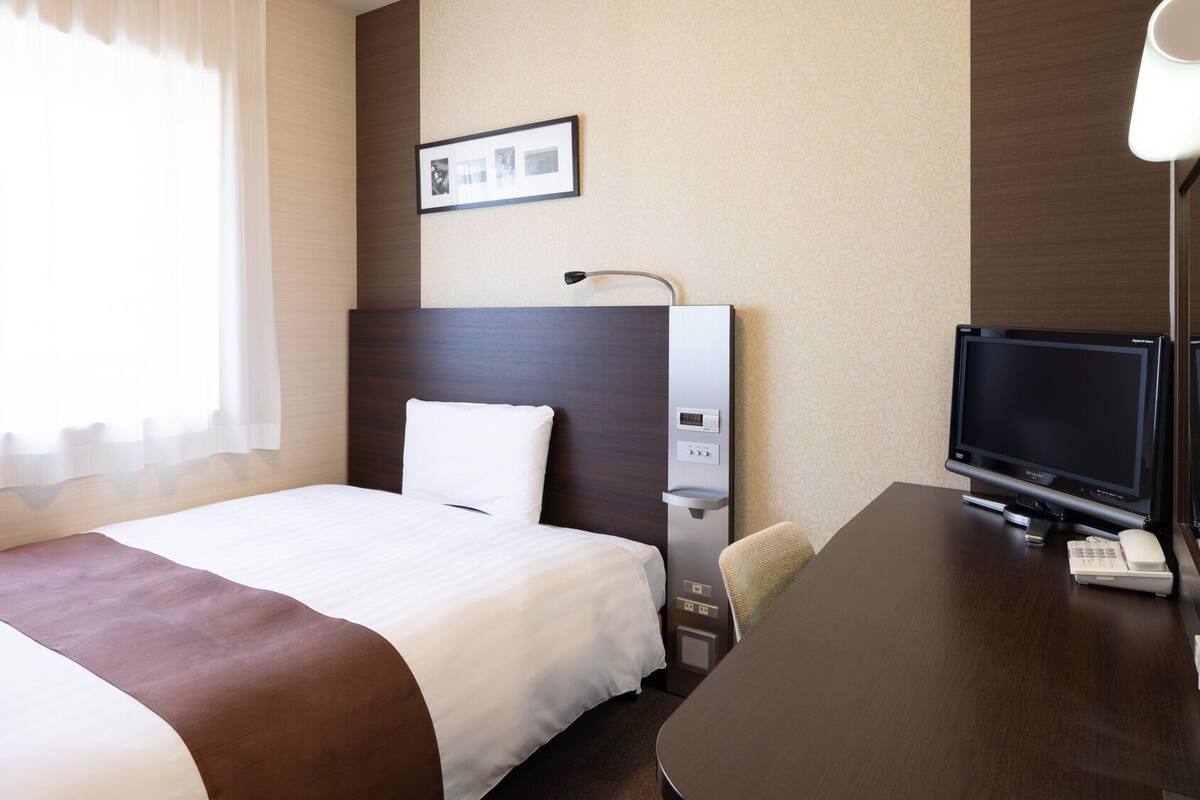 Een hotelkamer met een bed, een bureau met een televisie, een bureaulamp, een aan de muur hangend schilderij en een raam met gordijnen.