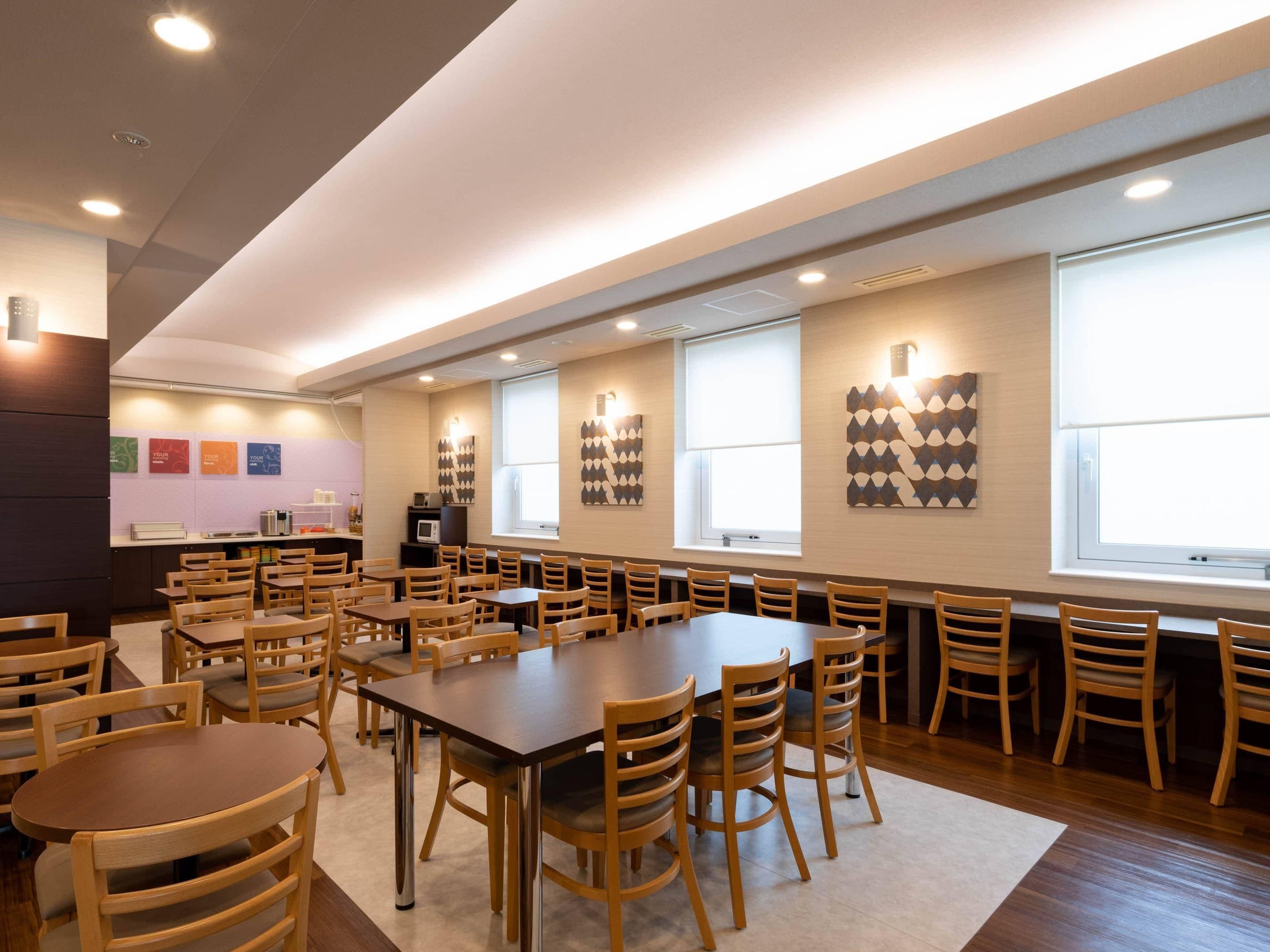Un salón de conferencias con varias mesas y sillas, una kitchenette y amplias ventanas.