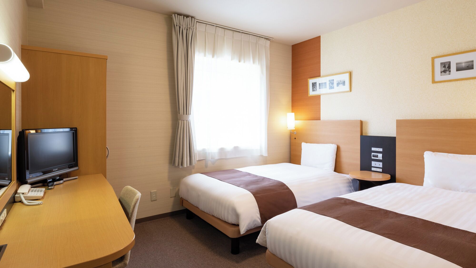 A hotel room with two beds, a desk with a computer and telephone, a window with curtains, and a wall-mounted light.