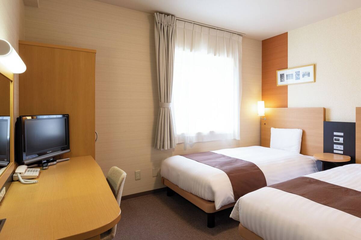 Een hotelkamer met twee bedden, een bureau met een computer en telefoon, een raam met gordijnen en een wandlamp.
