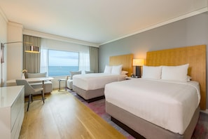 A hotel room with two beds, a desk, a chair, a lamp, and a view of the sea.