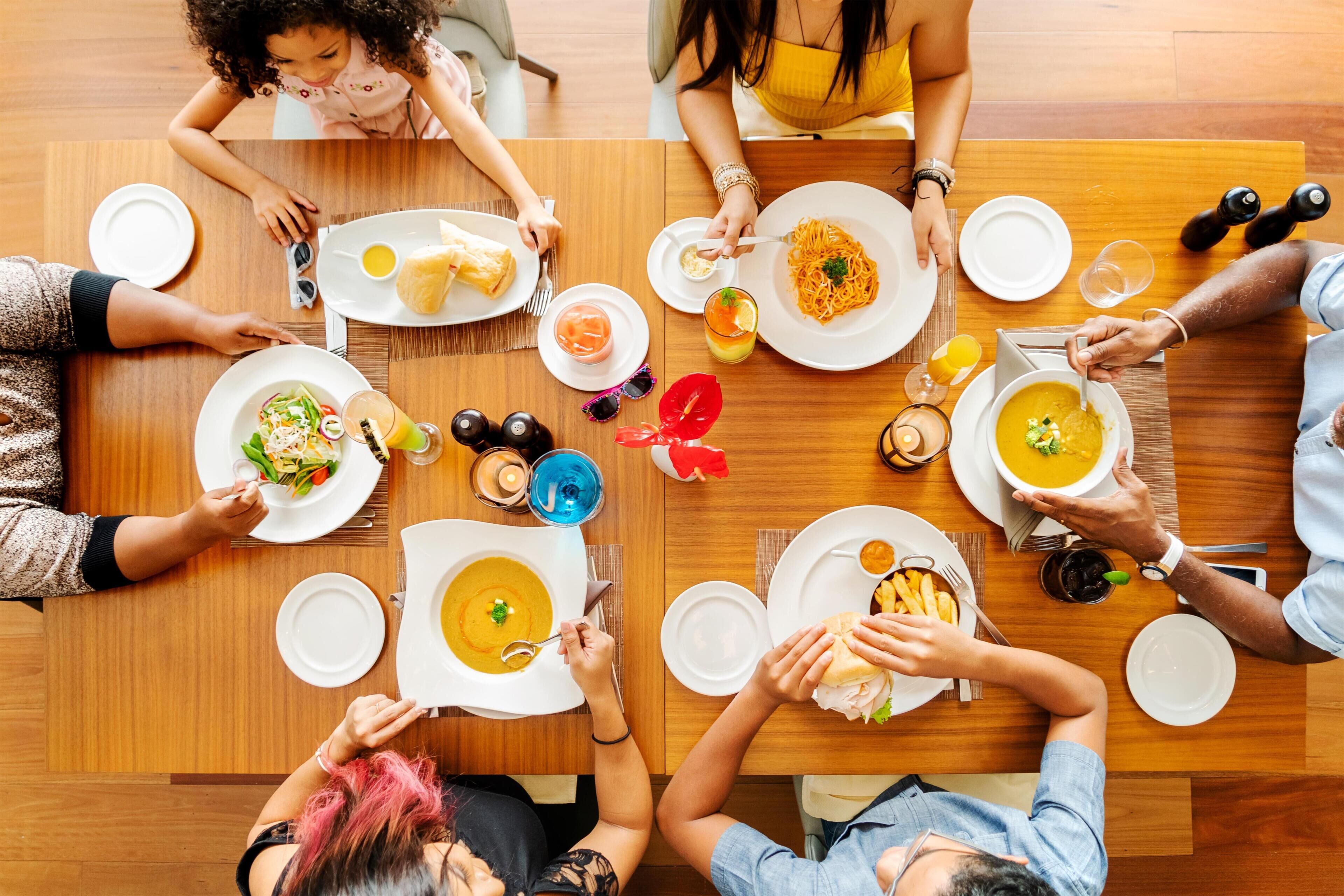 Um grupo de pessoas jantando em uma mesa de madeira com diversos pratos, incluindo sopa e massa, e temperos como saleiros e pimenteiros.