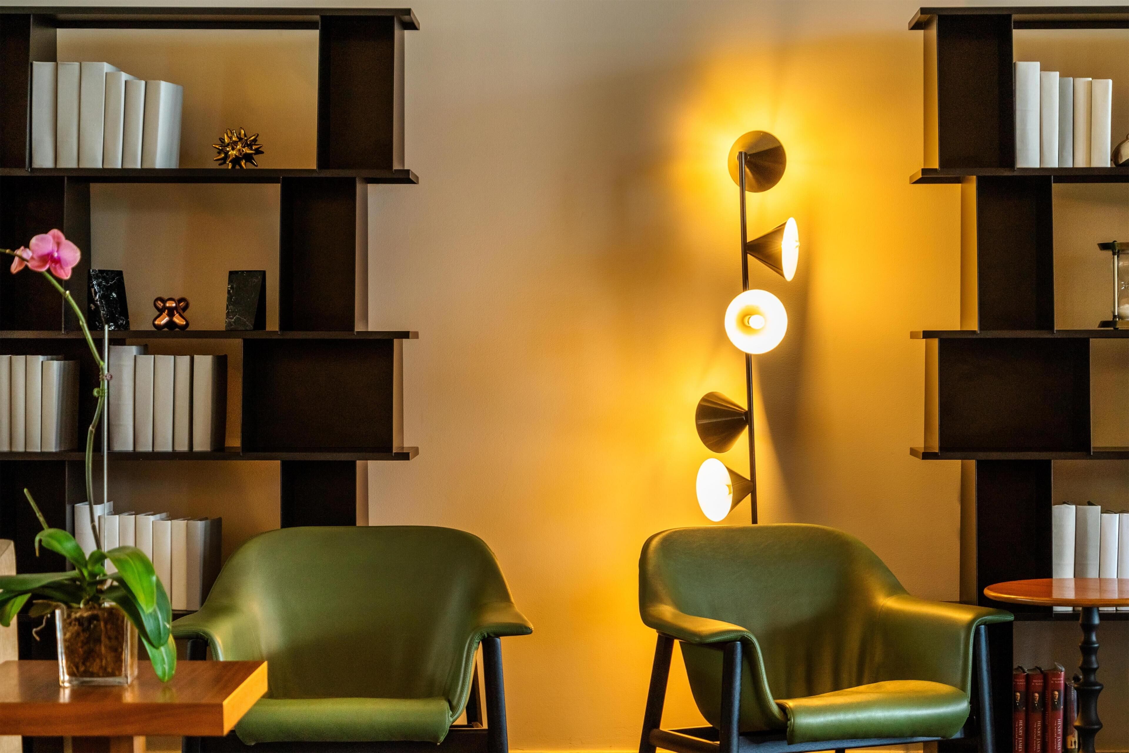 Quarto com duas cadeiras verdes, uma estante de livros e um aplique na parede.