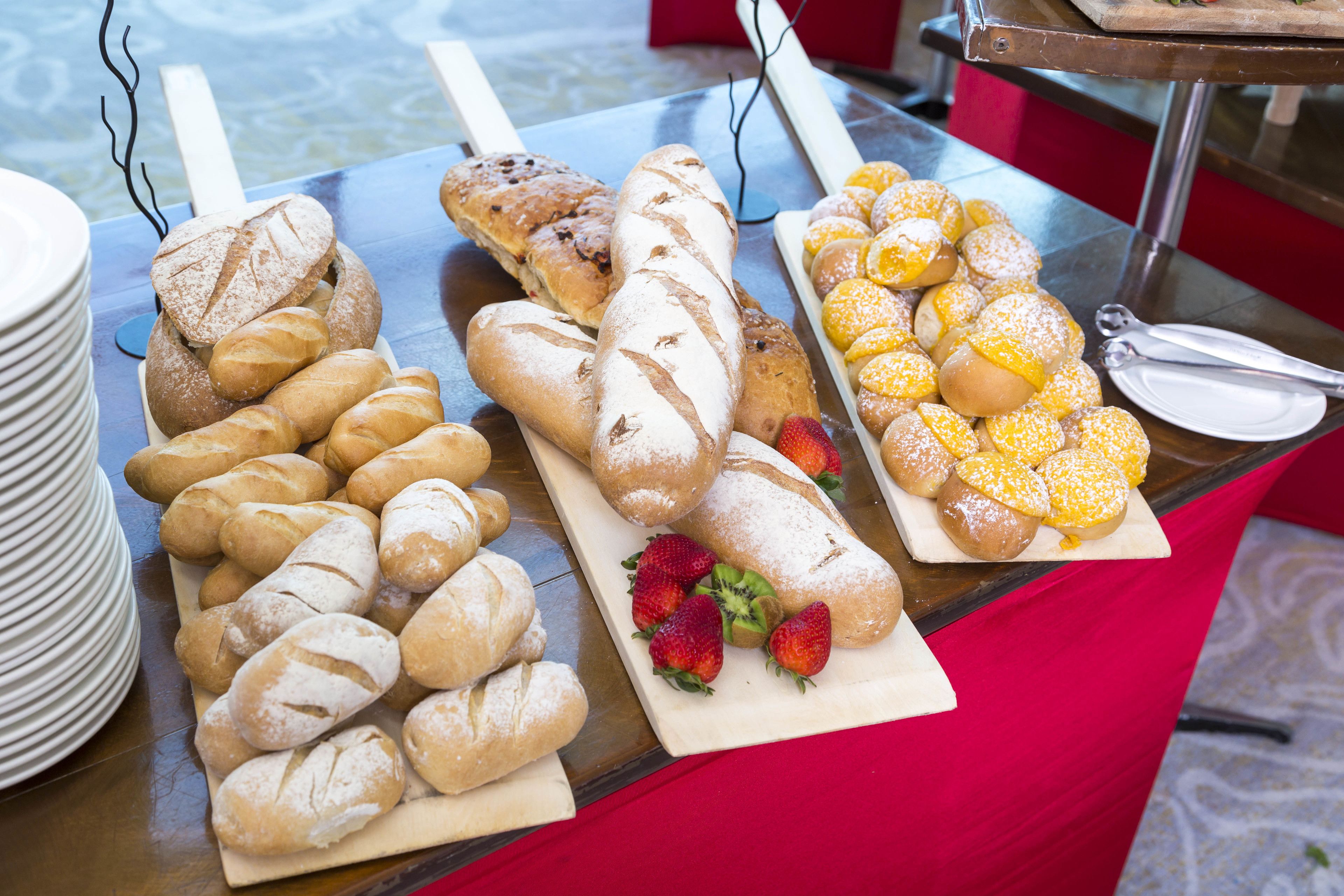 A buffet table with various breads and pastries, including croissants, a long loaf, and muffins dusted with powdered sugar.