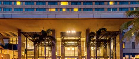 A hotel entrance with a covered outdoor area, palm trees, and a group of people sitting at tables.