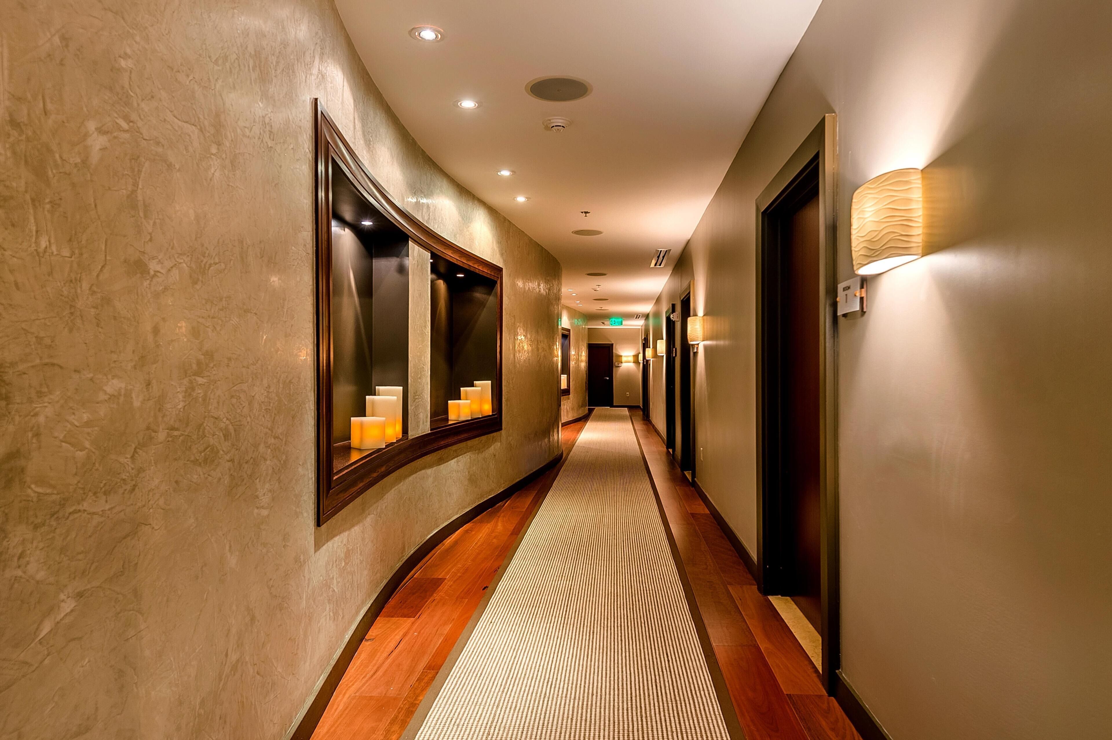 Un corridoio d'hotel con pavimento in legno, parete strutturata e un lampadario sulla destra.
