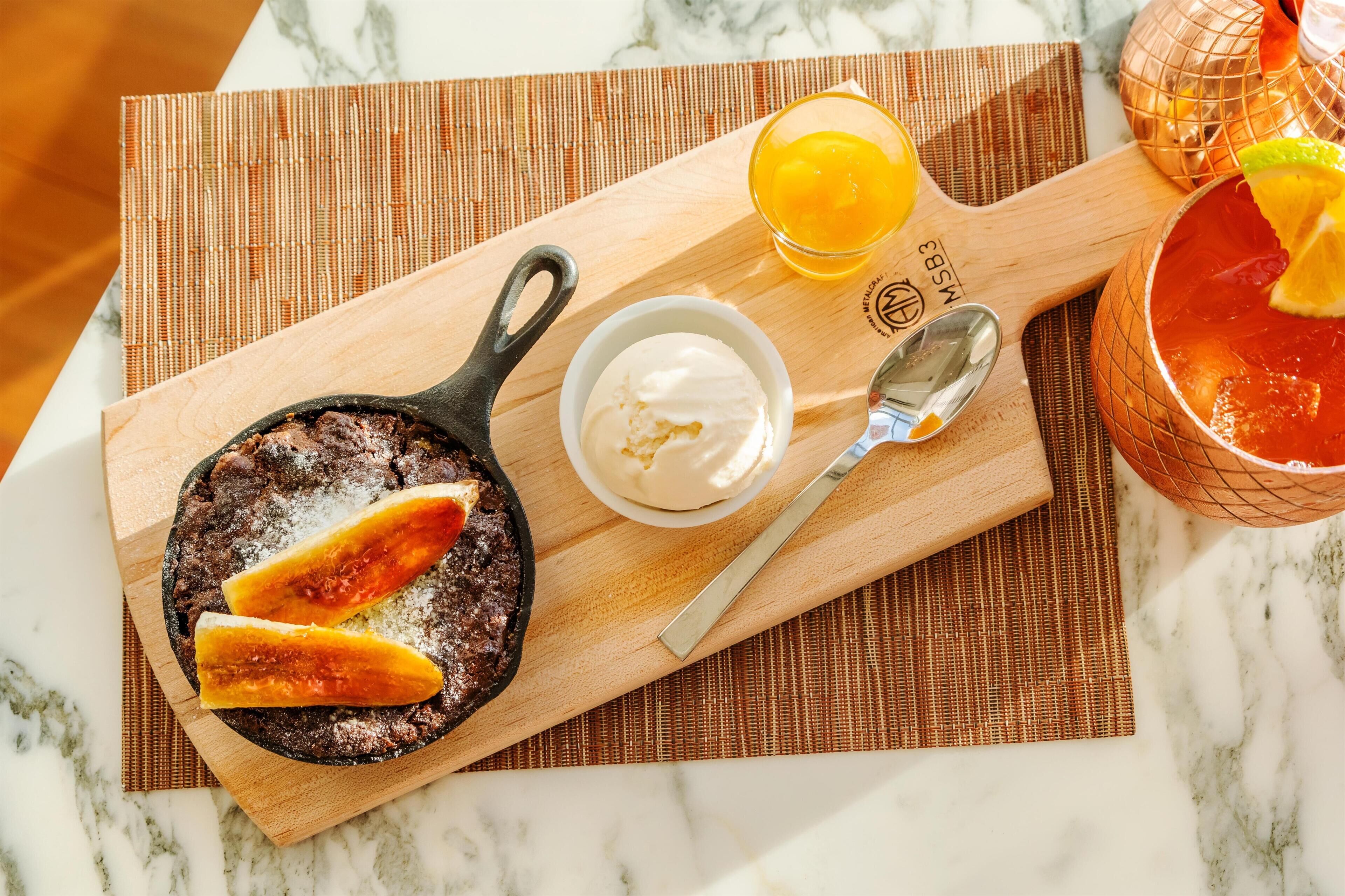 Un plato de postre con pastel de chocolate, crema batida en un bol y un vaso de jugo de naranja sobre una tabla de madera.