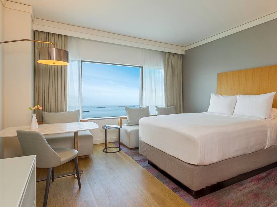 A hotel room with a large bed, a desk, a chair, a small table, and a view of the ocean.