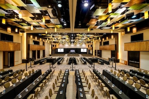 A conference room with rows of tables and chairs, a stage, and a ceiling with exposed beams and lighting fixtures.