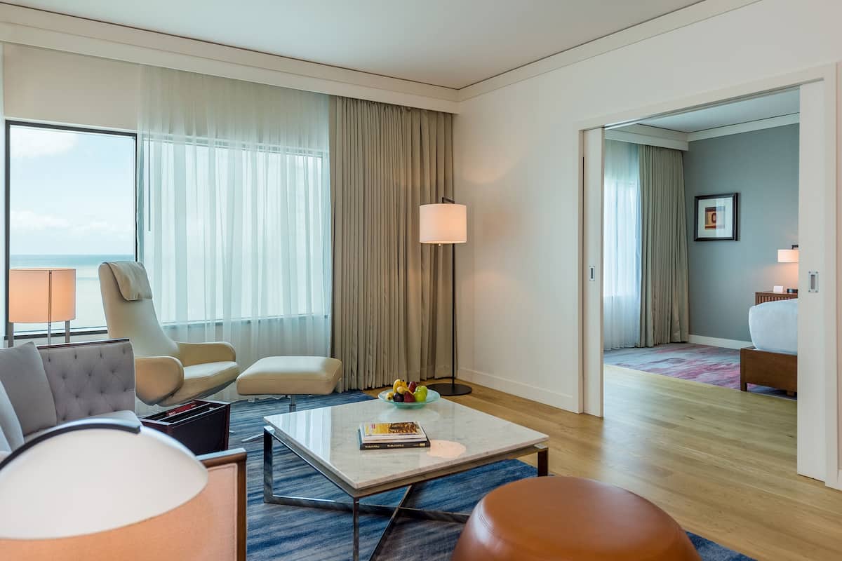 A hotel room with a large window, a sofa, a coffee table with books and a fruit bowl, a lamp, and a bed in the adjacent room.