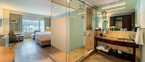 A modern hotel room with a glass-enclosed shower, a large bed, a desk, and a view of the city.