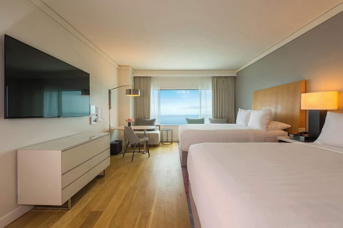 A hotel room with two beds, a desk, a chair, a TV, and a view of the sea.