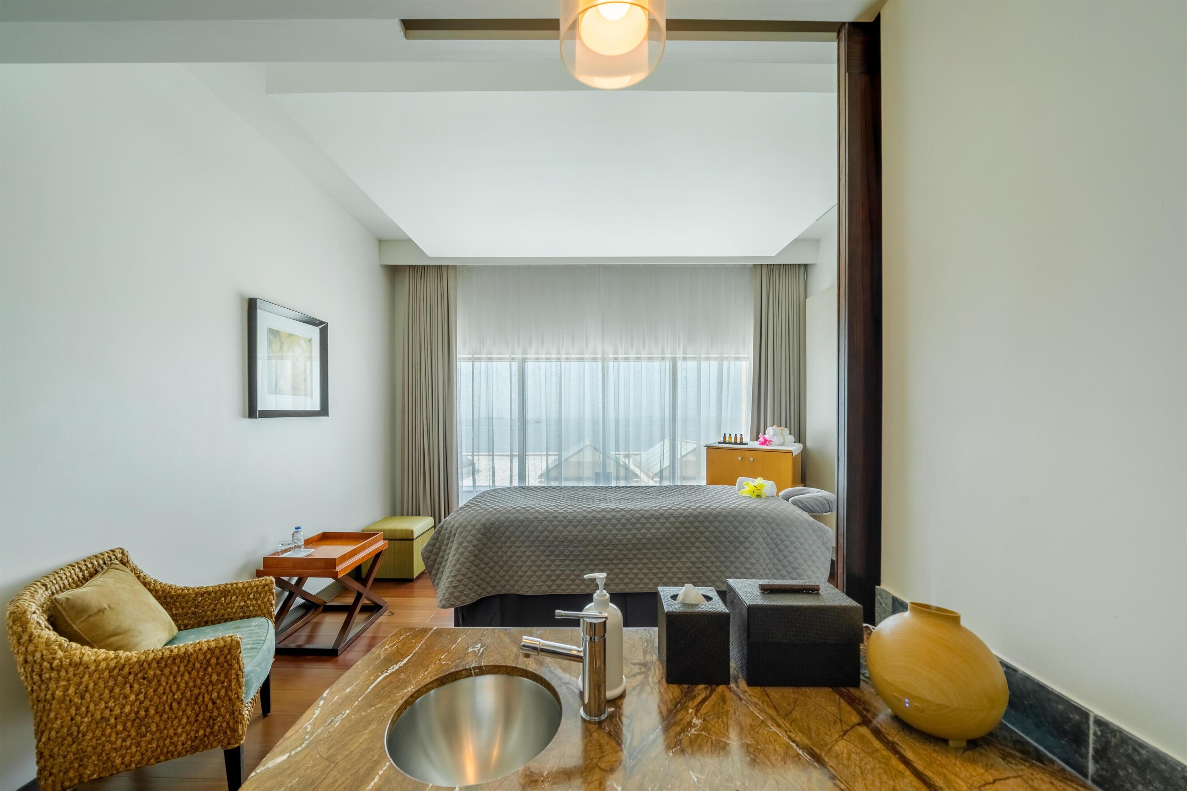 Une chambre d’hôtel avec un lit, une salle de bain comprenant un lavabo et un miroir, une chaise en osier, une petite table et un grand vase.
