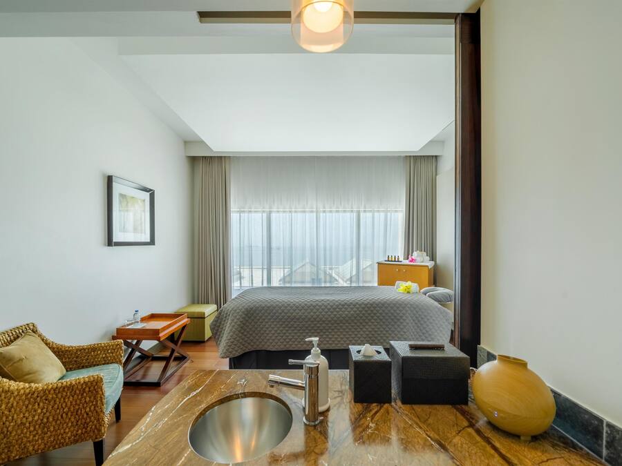 Une chambre d’hôtel avec un lit, une salle de bain comprenant un lavabo et un miroir, une chaise en osier, une petite table et un grand vase.