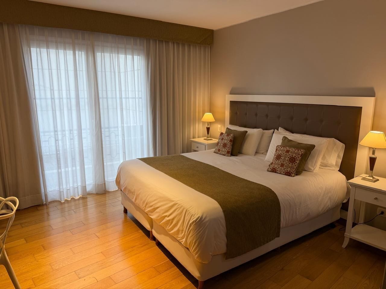 Een net opgemaakt bed met hoofdbord, twee nachtkastjes met lampen, en een groot raam met dunne gordijnen.