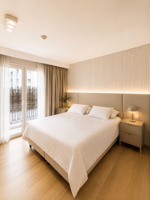 Una habitación de hotel con una cama grande, mesitas de noche, una mesita con lámpara, una ventana con cortinas y vistas al exterior.