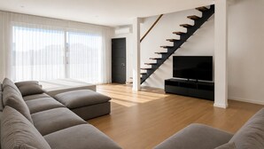 Een moderne woonkamer met een lange grijze bank, houten vloeren, een zwarte trap en een tv-meubel.