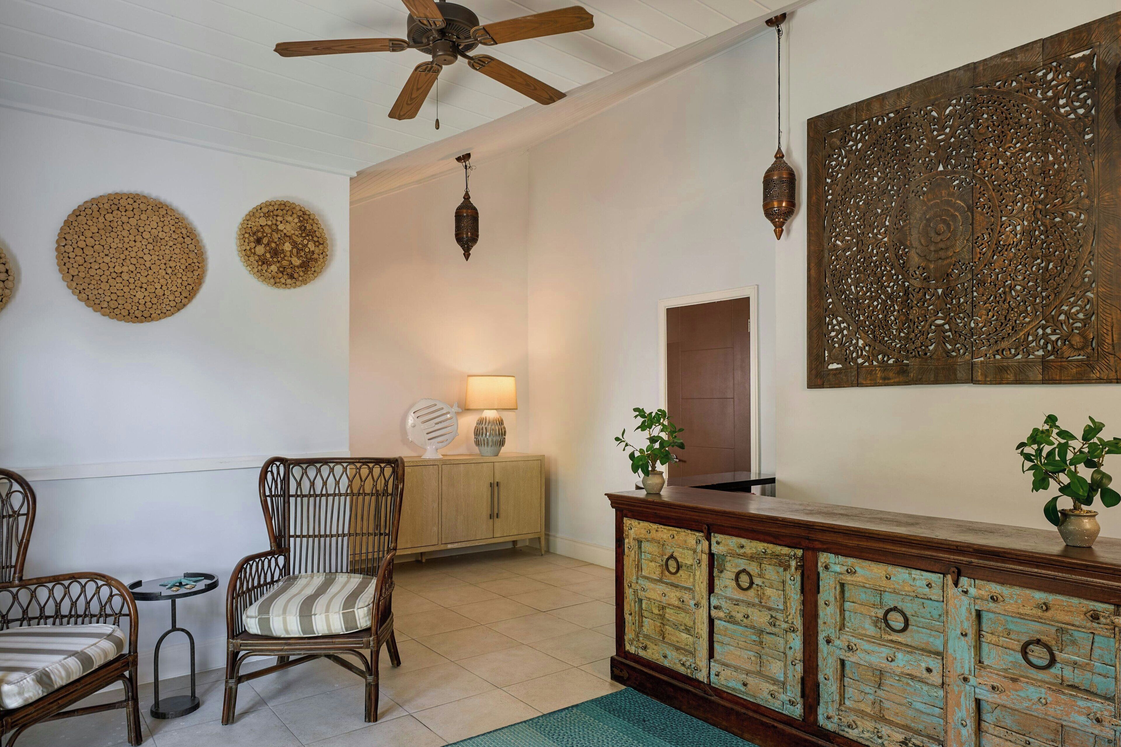 Habitació amb cadires de vimetla, un armari de fusta amb calaixos, un ventilador de sostre i una peça decorativa a la paret.