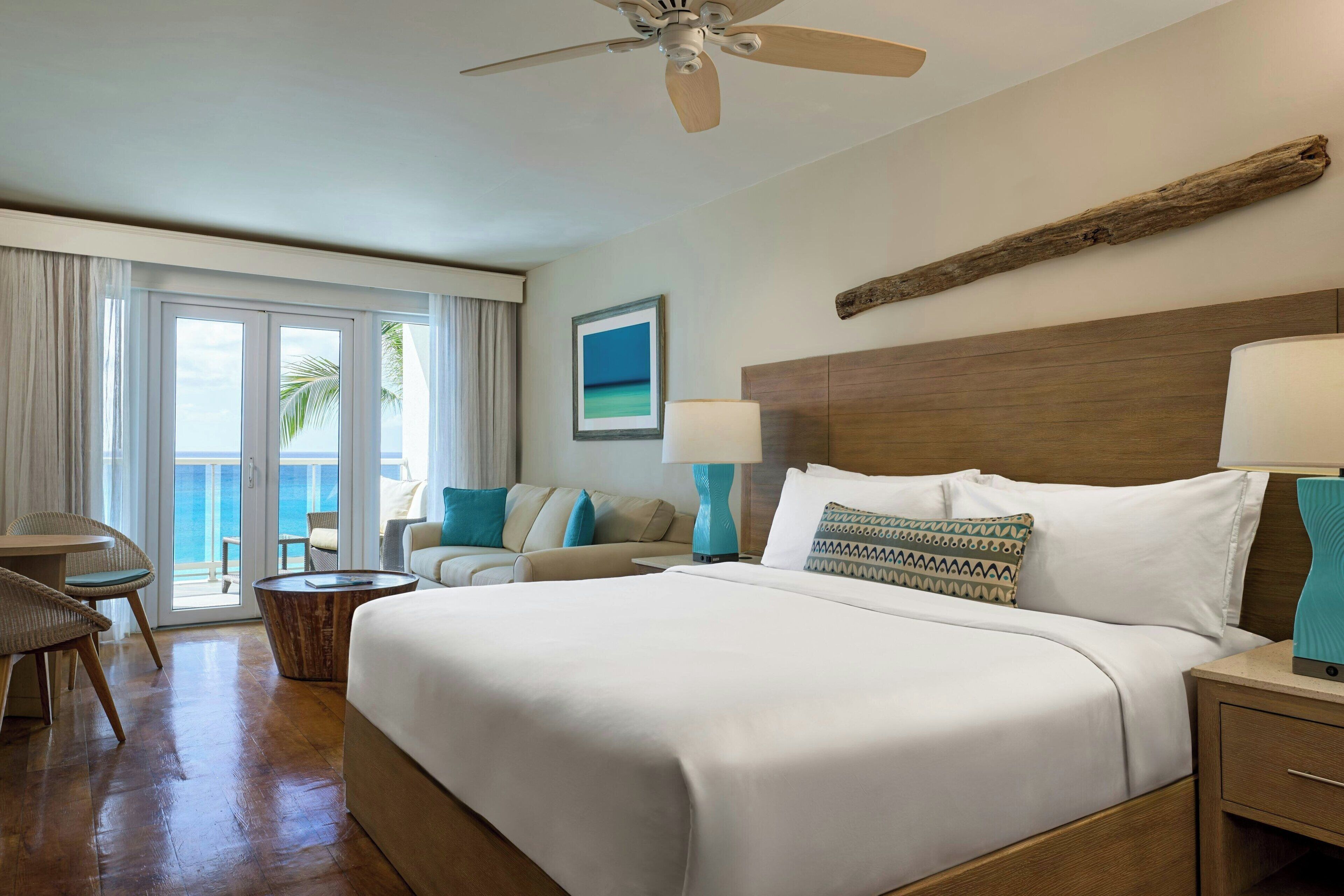 A hotel room with a large bed, a ceiling fan, a wooden headboard, a view of the ocean through the balcony doors, and a wooden side table with a lamp.