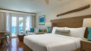 Habitación de hotel con una cama grande, un ventilador de techo, un cabecero de madera, vista al océano a través de las puertas del balcón y una mesita de noche de madera con una lámpara.