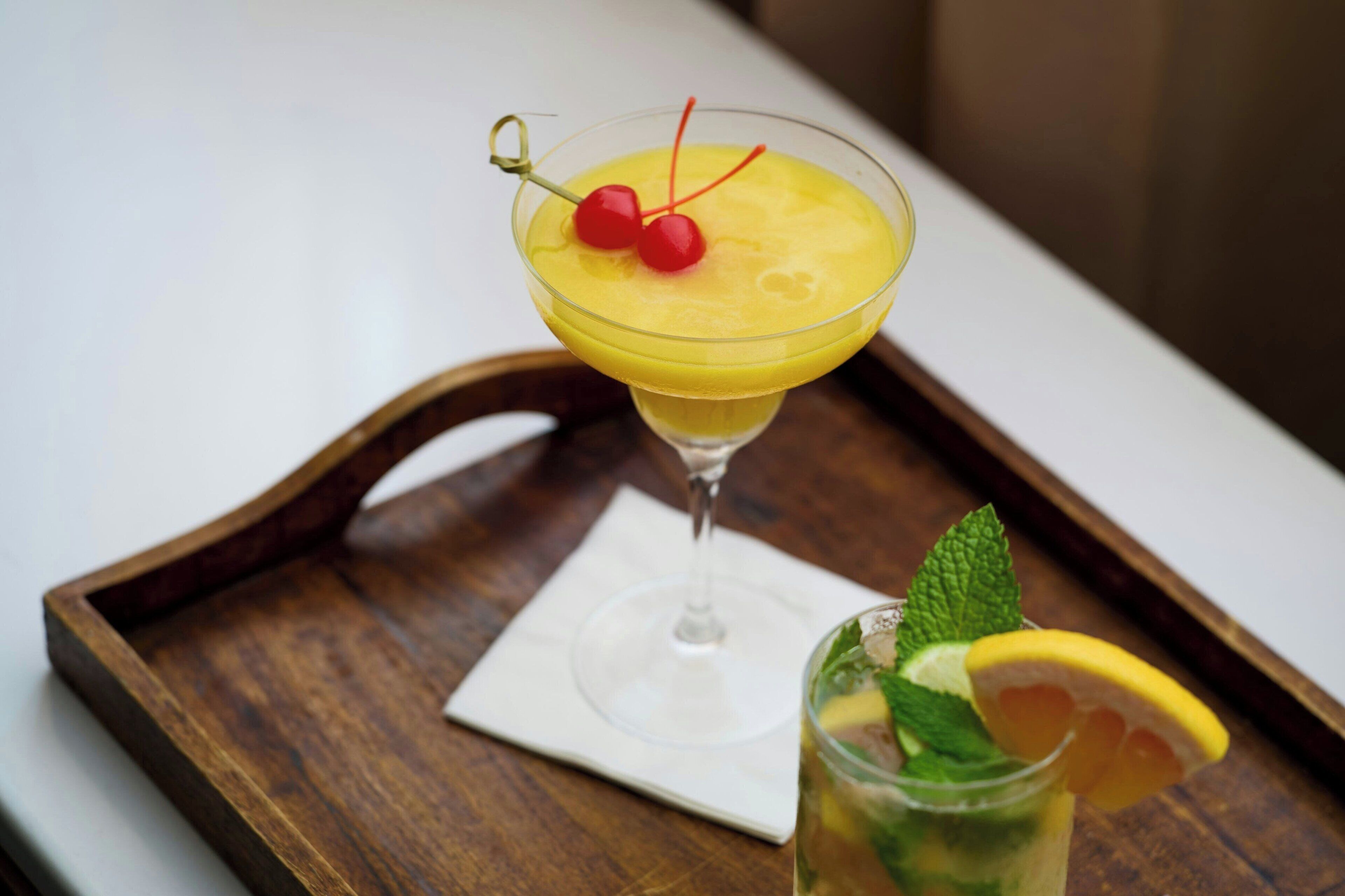 A cocktail in a martini glass with a cherry garnish on a wooden tray.